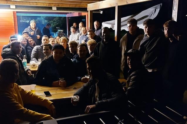 Gruppe von M&auml;nnern sitzt an einem Tisch unter einem &uuml;berdachten Bereich bei Nacht, mit Getr&auml;nken und einer warmen Atmosph&auml;re.