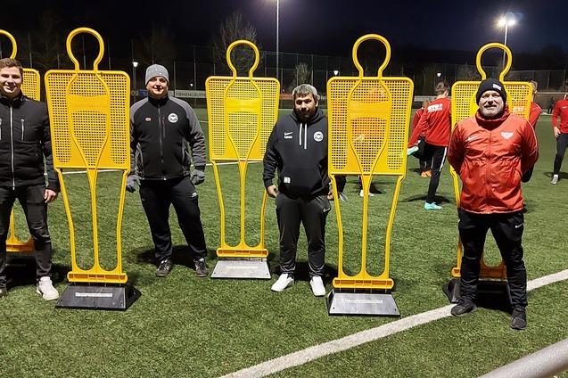 Training auf dem Fu&szlig;ballplatz mit vier orangefarbenen Trainingshilfen und drei Trainern in Sportkleidung.