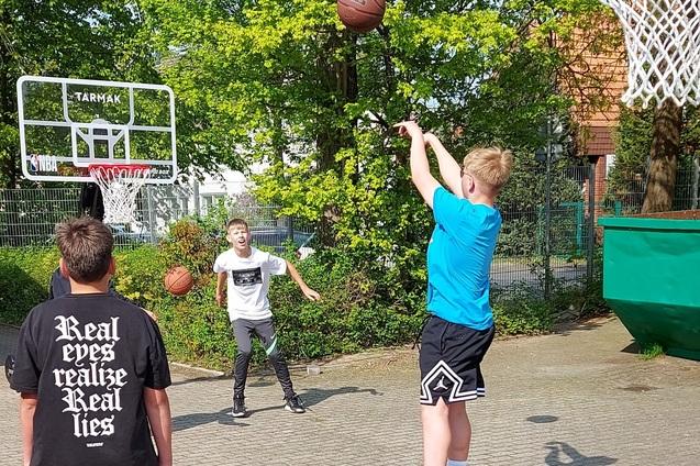 Junger Spieler wirft Basketball zum Korb, w&auml;hrend andere Spieler im Hintergrund warten, auf einem Freiplatz.