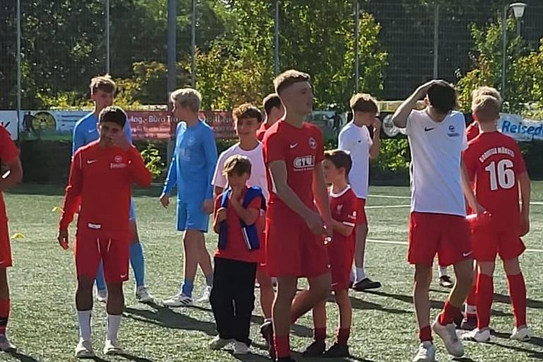 Mehrere Jugendliche in roten Fu&szlig;balltrikots stehen auf einem Kunstrasenplatz. Einige sind in Gruppen versammelt.