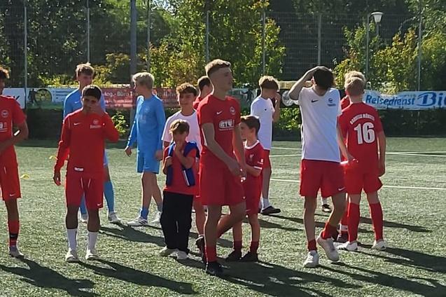Mehrere Jugendliche in roten Fu&szlig;balltrikots stehen auf einem Kunstrasenplatz. Einige sind in Gruppen versammelt.