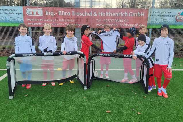 Gruppe von Kindern in Fu&szlig;balltrikots steht an einem Kunstrasenplatz mit Minitoren.