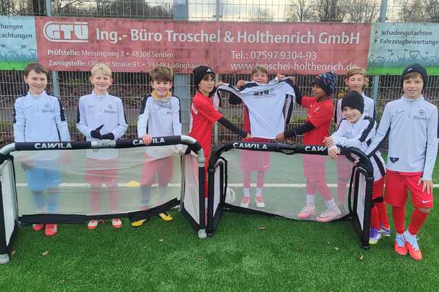 Gruppe von Kindern in Fu&szlig;balltrikots steht an einem Kunstrasenplatz mit Minitoren.