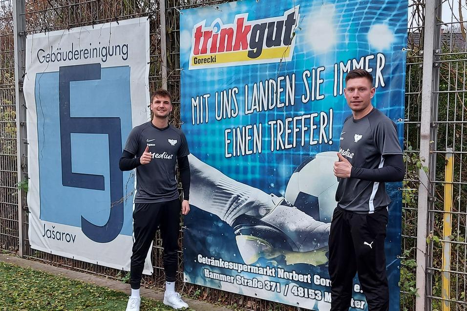Zwei Fu&szlig;ballspieler stehen daneben einem Werbebanner auf einem Sportplatz und geben Daumen hoch.