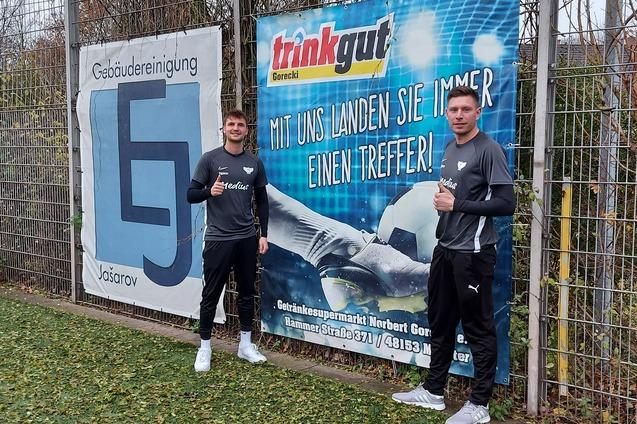 Zwei Fu&szlig;ballspieler stehen daneben einem Werbebanner auf einem Sportplatz und geben Daumen hoch.