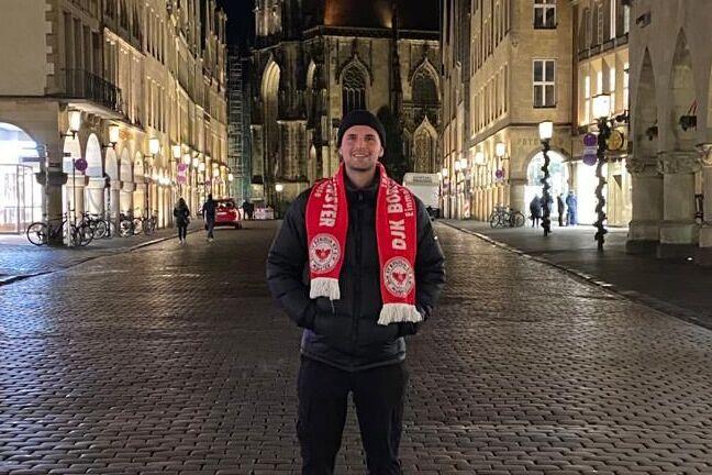 Junger Mann in Winterjacke und Schal steht nachts auf einer gepflasterten Stra&szlig;e vor einer Kirche und historischen Geb&auml;uden.