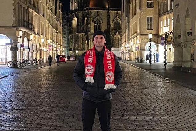 Junger Mann in Winterjacke und Schal steht nachts auf einer gepflasterten Stra&szlig;e vor einer Kirche und historischen Geb&auml;uden.