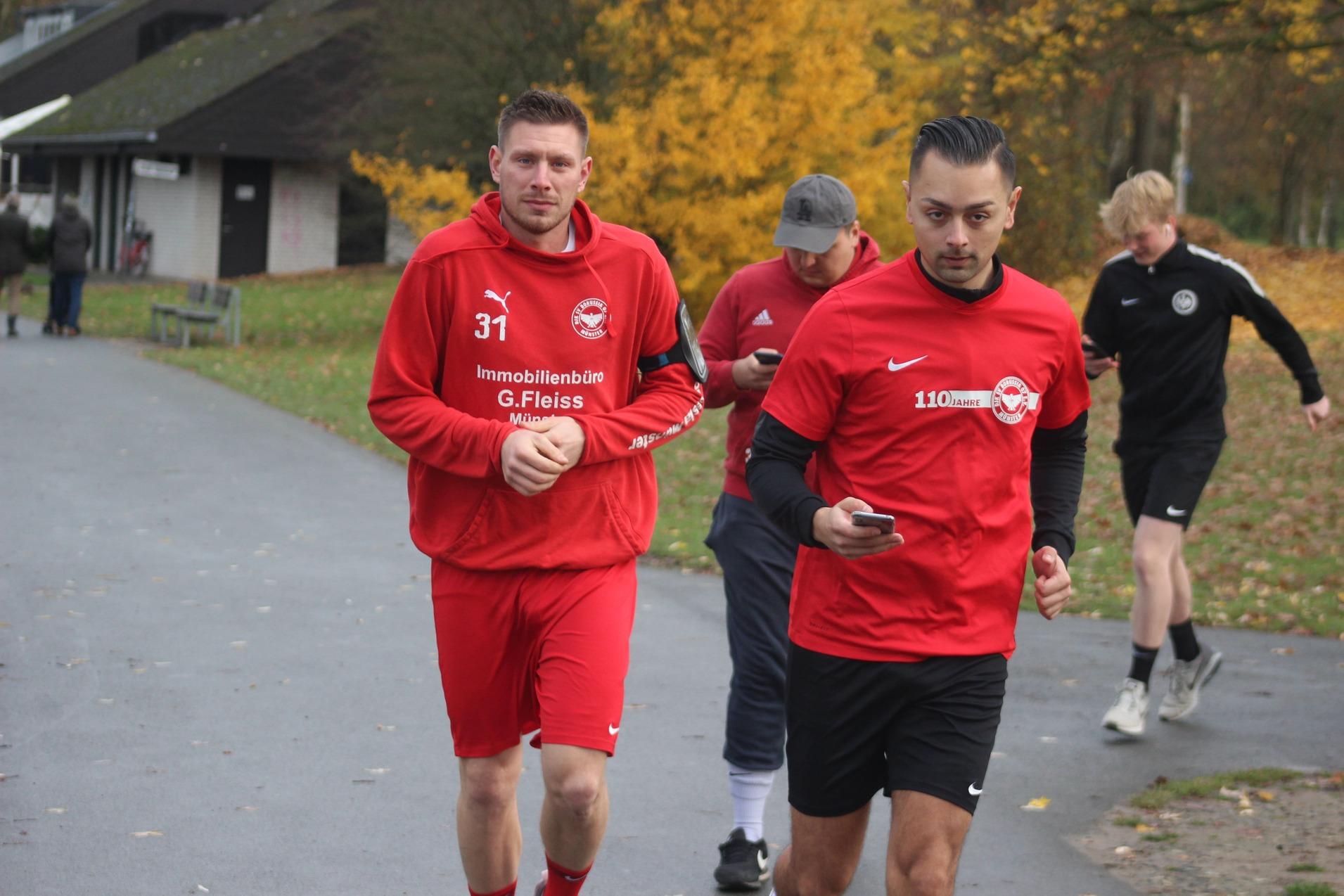 Zwei Sportler in roten Trainingsanz&uuml;gen joggen in einem Park, w&auml;hrend ein weiterer L&auml;ufer im Hintergrund vorbeil&auml;uft.
