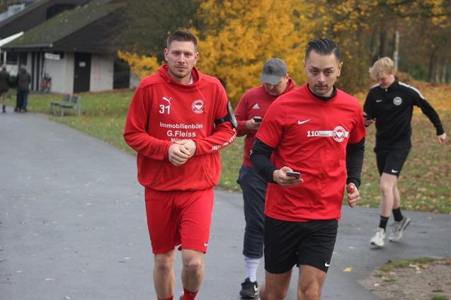 Zwei Sportler in roten Trainingsanz&uuml;gen joggen in einem Park, w&auml;hrend ein weiterer L&auml;ufer im Hintergrund vorbeil&auml;uft.