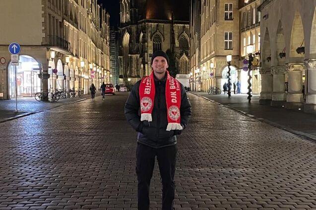 Junger Mann in Winterjacke und Schal steht nachts auf einer gepflasterten Stra&szlig;e vor einer Kirche und historischen Geb&auml;uden.