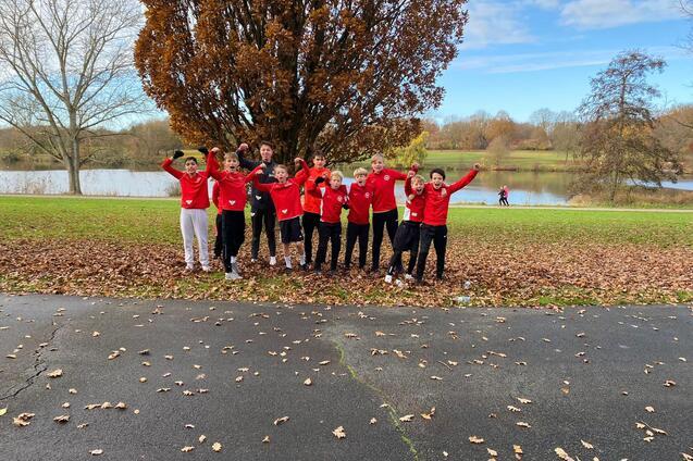 Gruppe von neun Personen in roten Jacken posiert freudig vor einem See mit Herbstlaub und B&auml;umen im Hintergrund.