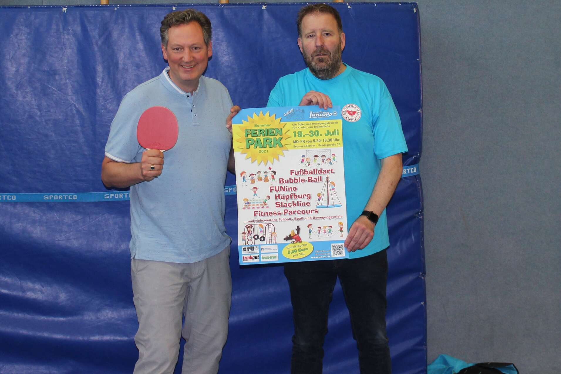 Zwei M&auml;nner, einer mit Tischtennisschl&auml;ger, pr&auml;sentieren ein Plakat in einer Sporthalle.
