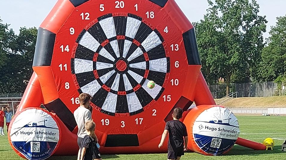 Kinder kicken B&auml;lle auf ein gro&szlig;es, aufblasbares Dartboard auf einem Rasenplatz. Zwei Fu&szlig;ballb&auml;lle liegen auf dem Boden.
