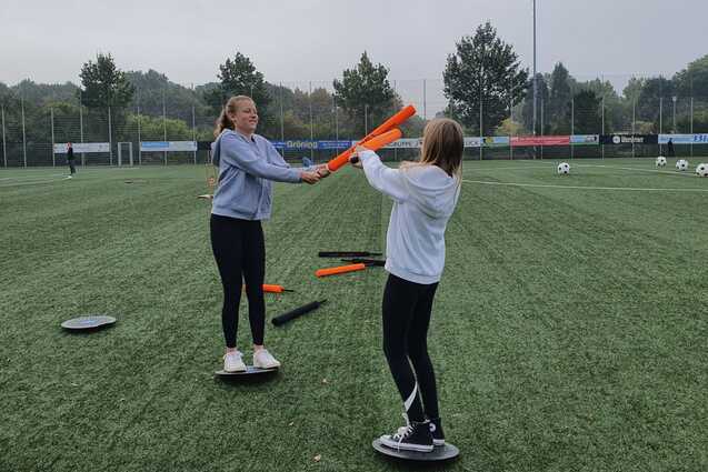 Zwei M&auml;dchen trainieren auf einem Sportplatz und balancieren auf runden Plattformen mit bunten St&auml;ben in der Hand.