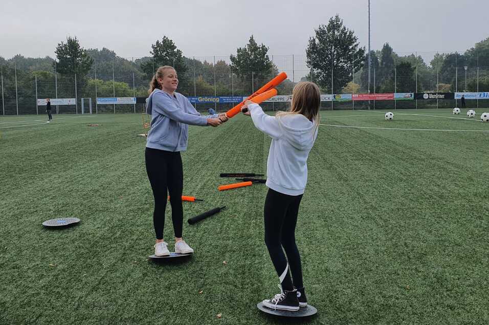 Zwei M&auml;dchen trainieren auf einem Sportplatz und balancieren auf runden Plattformen mit bunten St&auml;ben in der Hand.
