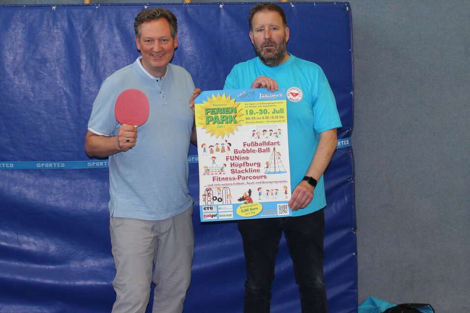 Zwei M&auml;nner, einer mit Tischtennisschl&auml;ger, pr&auml;sentieren ein Plakat in einer Sporthalle.