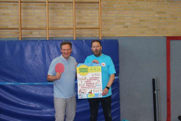 Zwei M&auml;nner, einer mit Tischtennisschl&auml;ger, pr&auml;sentieren ein Plakat in einer Sporthalle.