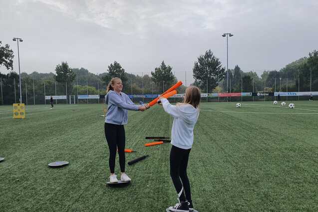 Zwei M&auml;dchen trainieren auf einem Sportplatz und balancieren auf runden Plattformen mit bunten St&auml;ben in der Hand.
