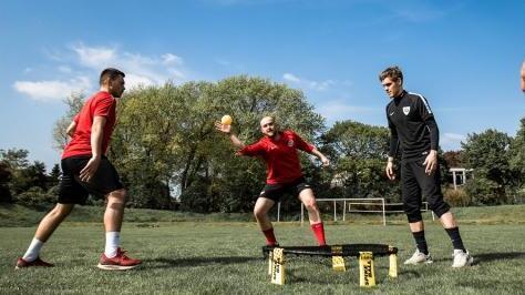 Vier Fu&szlig;ballspieler trainieren auf einem Rasenplatz, w&auml;hrend einer einen Ball zu einem Mitspieler wirft.