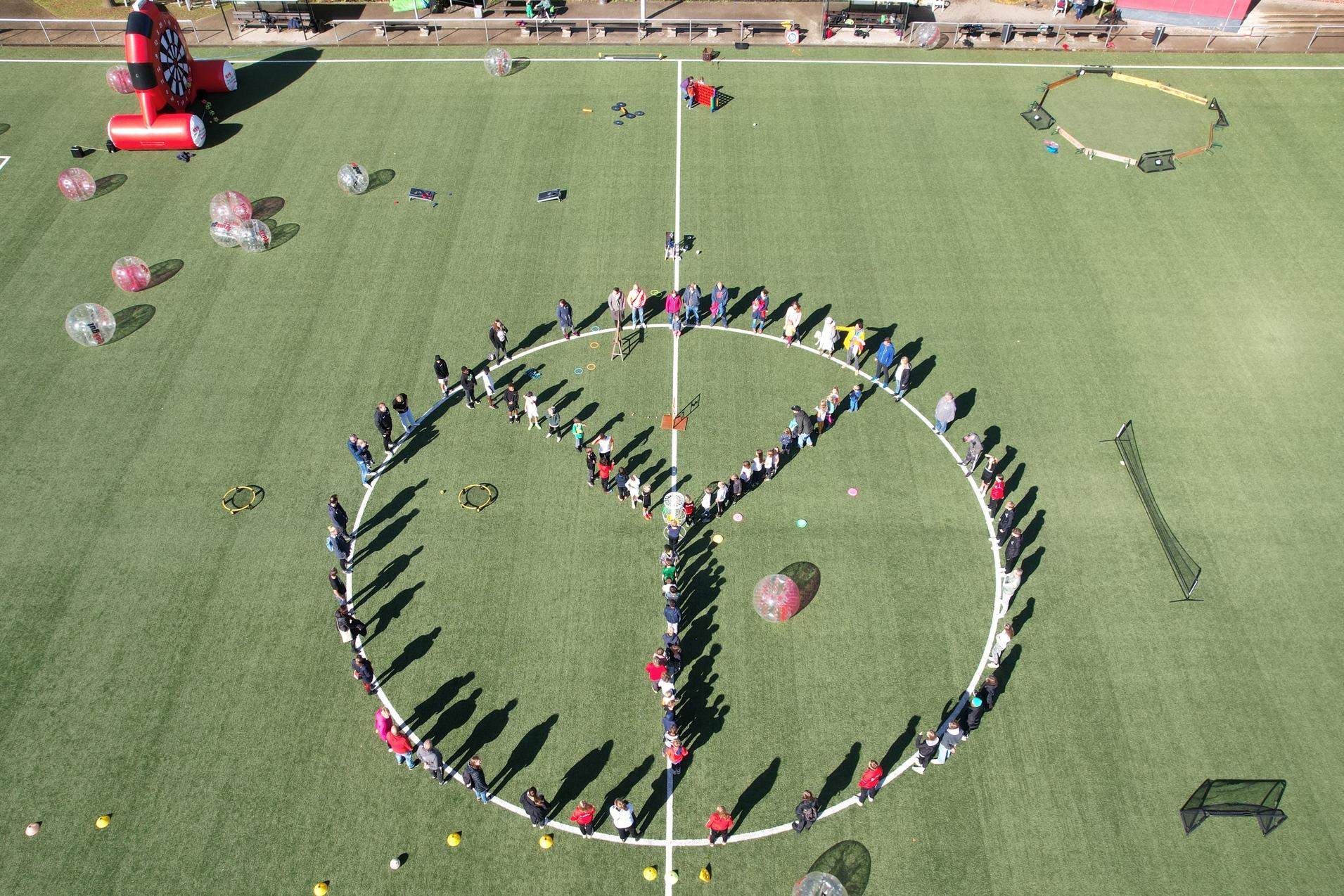 Menschen bilden einen gro&szlig;en Kreis mit einem Peace-Zeichen auf einem Fu&szlig;ballfeld, umgeben von bunten Ballons und Equipment.