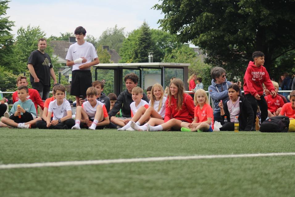 Gruppe von Kindern und Erwachsenen sitzend am Rande eines Fu&szlig;ballfeldes, umgeben von gr&uuml;ner Natur.