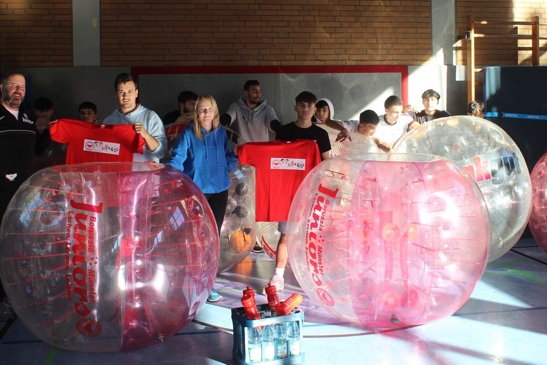 Gruppenspieler in aufblasbaren Bubbles halten T-Shirts und stehen in einer Sporthalle neben Wasserflaschen und Basketballkorb.