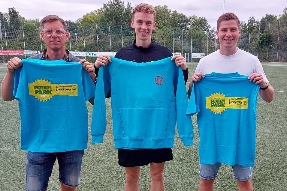 Drei M&auml;nner stehen auf einem Fu&szlig;ballplatz und halten leuchtend blaue Sweatshirts mit einem bunten Logo in den H&auml;nden.