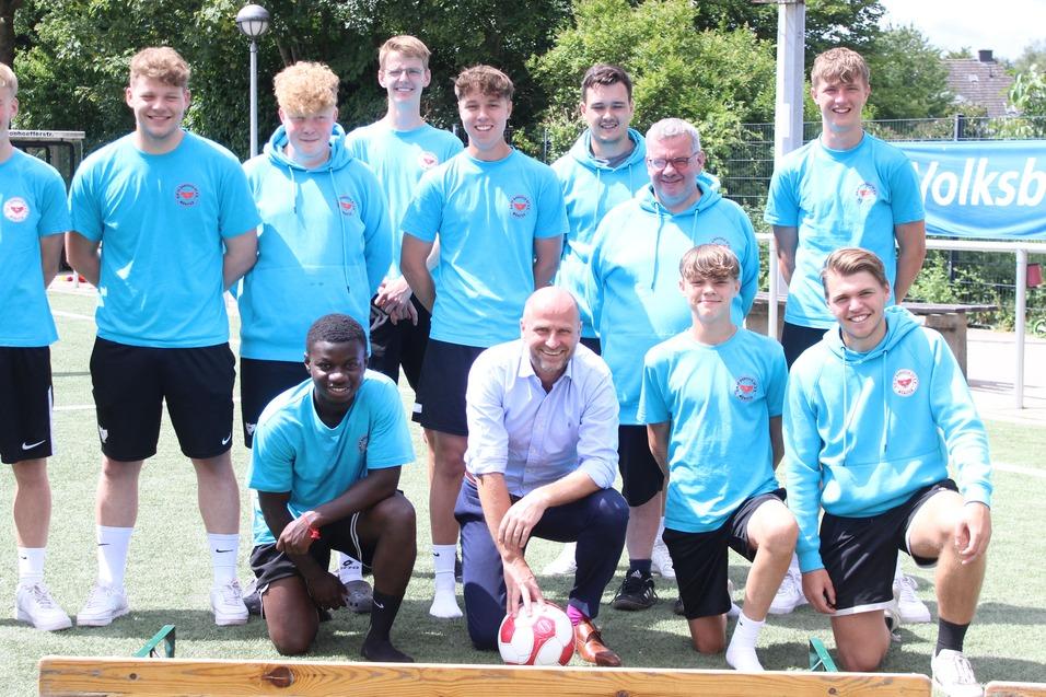 Gruppenfoto von zehn Fu&szlig;ballspielern in blauen Shirts und zwei Betreuern auf einem Sportplatz mit Banner im Hintergrund.