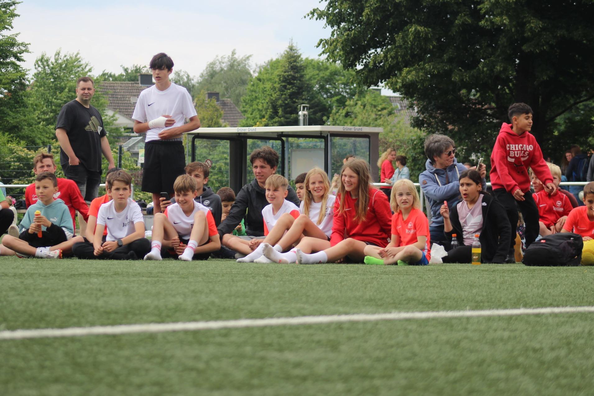 Gruppe von Kindern und Erwachsenen sitzend am Rande eines Fu&szlig;ballfeldes, umgeben von gr&uuml;ner Natur.