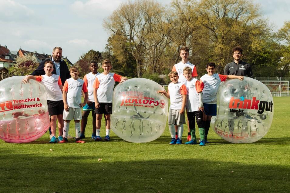 Gruppe von Kindern und Erwachsenen in Zorb-B&auml;llen auf einem Fu&szlig;ballfeld, umgeben von B&auml;umen und einem Sportplatz.