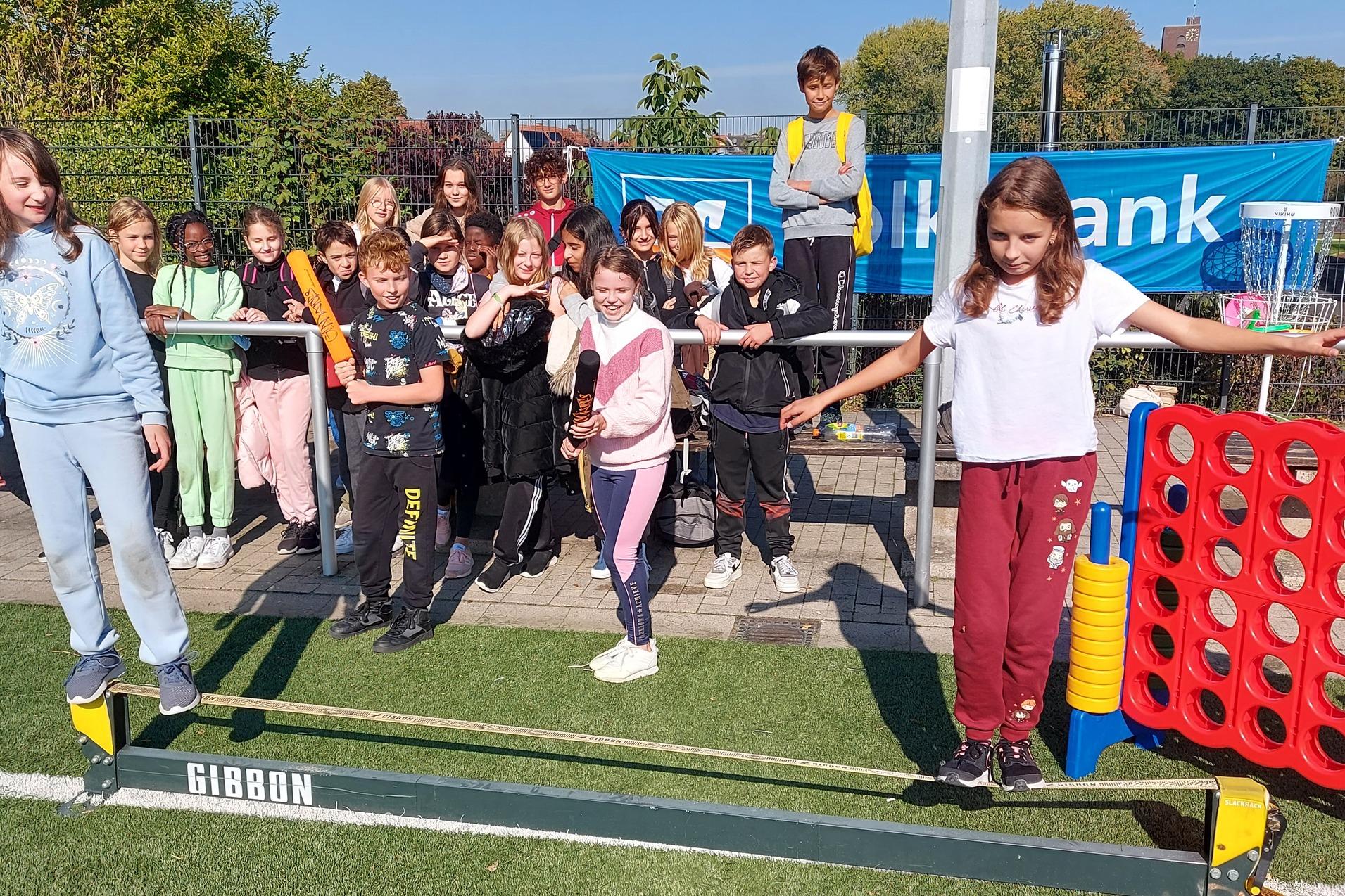 Gruppe von Kindern steht auf einer Slackline im Freien, einige lachen, w&auml;hrend andere zuschauen.