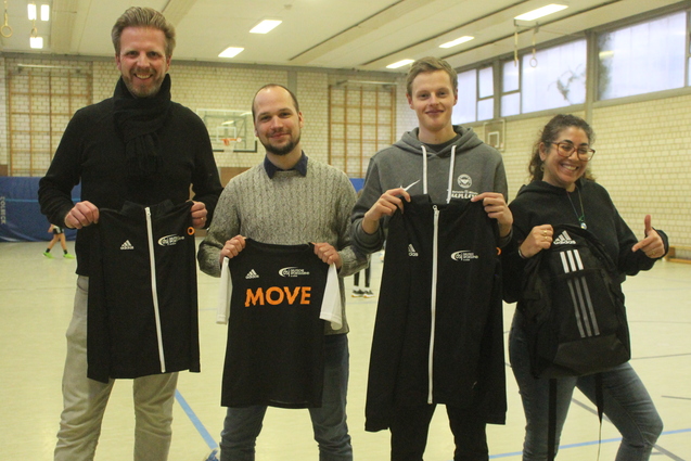 Vier Personen in Sportkleidung halten T-Shirts mit dem Schriftzug &bdquo;MOVE&ldquo; in einer Sporthalle.