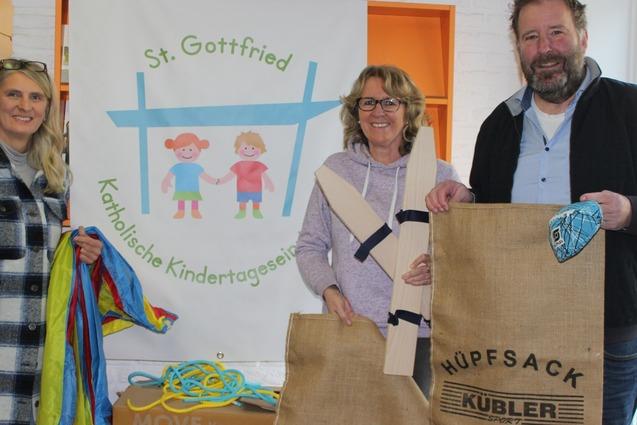 Drei Personen stehen vor einem Banner der katholischen Kindertagesst&auml;tte St. Gottfried und halten Aktivit&auml;tss&auml;cke.