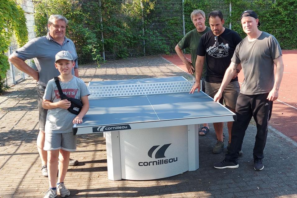 Vier Personen, darunter ein Kind, posieren um einen Tischtennistisch auf einem Au&szlig;enplatz. Hintergrund mit Tor und B&auml;umen.