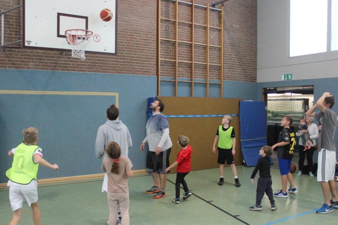 Gruppe von Kindern und Jugendlichen spielt Basketball in einer Sporthalle, ein Spieler wirft den Ball zum Korb.