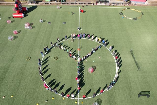 Menschen bilden einen gro&szlig;en Kreis mit einem Peace-Zeichen auf einem Fu&szlig;ballfeld, umgeben von bunten Ballons und Equipment.