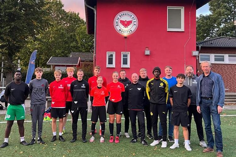 Gruppe von Teenagern in Sportkleidung steht vor einem Vereinsgeb&auml;ude auf einem Fu&szlig;ballplatz.