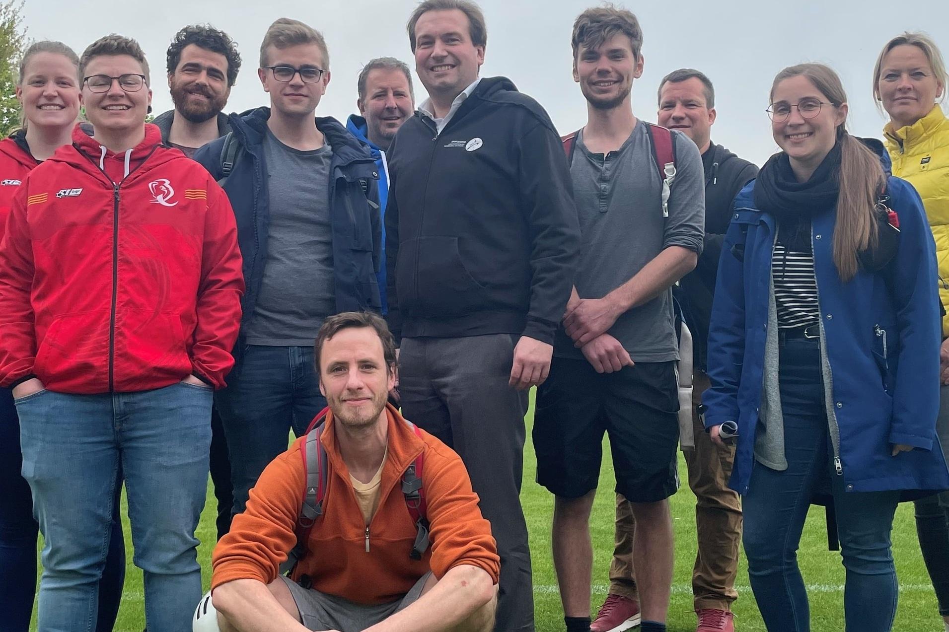 Gruppe von zehn Personen l&auml;chelt auf einem gr&uuml;nen Rasenplatz, einige tragen Sportjacken und stehen in lockerer Pose.