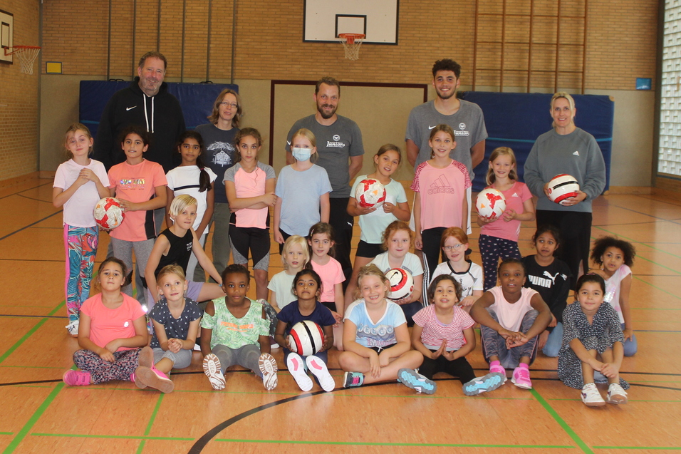 Gruppenfoto von Kindern und Trainern in einer Sporthalle, einige halten Volleyballs, umgeben von Holzfu&szlig;boden und W&auml;nden.