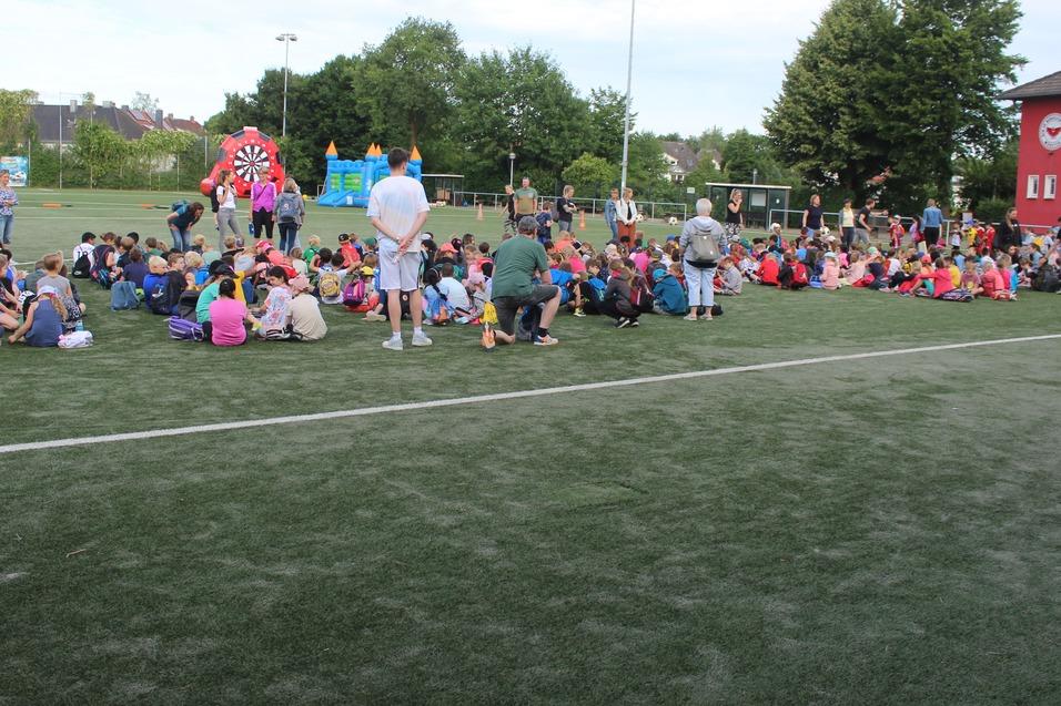 Viele Kinder sitzen auf einem Sportplatz, w&auml;hrend eine Veranstaltung mit bunten Attraktionen im Hintergrund stattfindet.