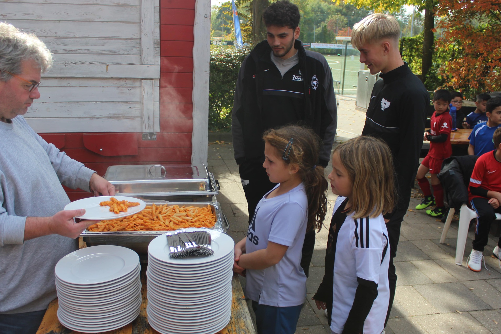 Ein Mann serviert Pasta an Kinder in Sportkleidung vor einem Tisch mit wei&szlig;en Tellern im Freien.