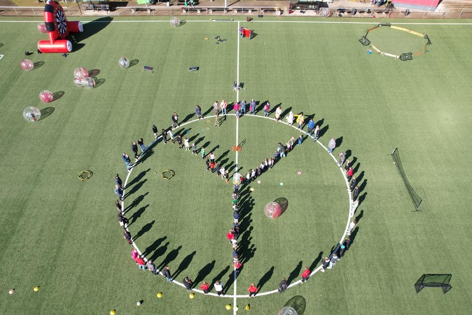 Menschen bilden einen gro&szlig;en Kreis mit einem Peace-Zeichen auf einem Fu&szlig;ballfeld, umgeben von bunten Ballons und Equipment.