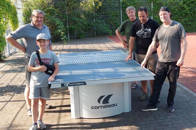 Vier Personen, darunter ein Kind, posieren um einen Tischtennistisch auf einem Au&szlig;enplatz. Hintergrund mit Tor und B&auml;umen.