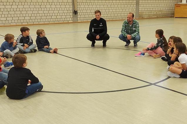 Zwei Erwachsene sitzen auf dem Boden, umgeben von einer Gruppe von Kindern in einer Sporthalle.