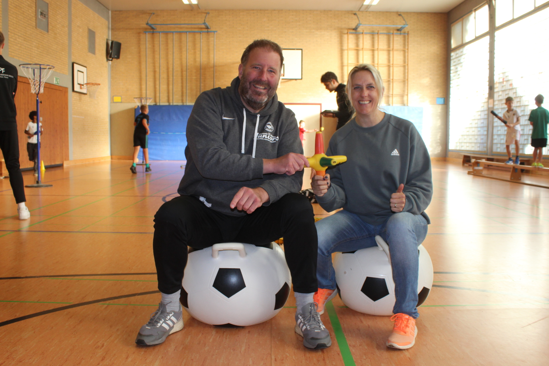 Zwei Personen sitzen auf gro&szlig;en, runden Fu&szlig;b&auml;llen in einer Sporthalle und l&auml;cheln in die Kamera.