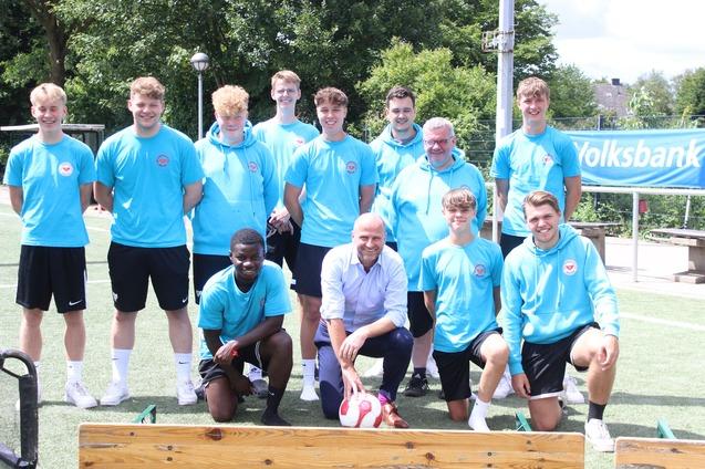 Gruppenfoto von zehn Fu&szlig;ballspielern in blauen Shirts und zwei Betreuern auf einem Sportplatz mit Banner im Hintergrund.