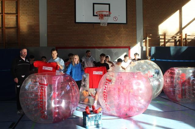 Gruppenspieler in aufblasbaren Bubbles halten T-Shirts und stehen in einer Sporthalle neben Wasserflaschen und Basketballkorb.