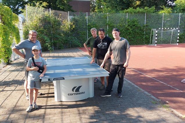 Vier Personen, darunter ein Kind, posieren um einen Tischtennistisch auf einem Au&szlig;enplatz. Hintergrund mit Tor und B&auml;umen.