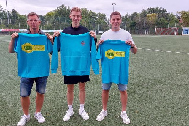 Drei M&auml;nner stehen auf einem Fu&szlig;ballplatz und halten leuchtend blaue Sweatshirts mit einem bunten Logo in den H&auml;nden.