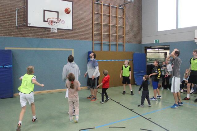 Gruppe von Kindern und Jugendlichen spielt Basketball in einer Sporthalle, ein Spieler wirft den Ball zum Korb.